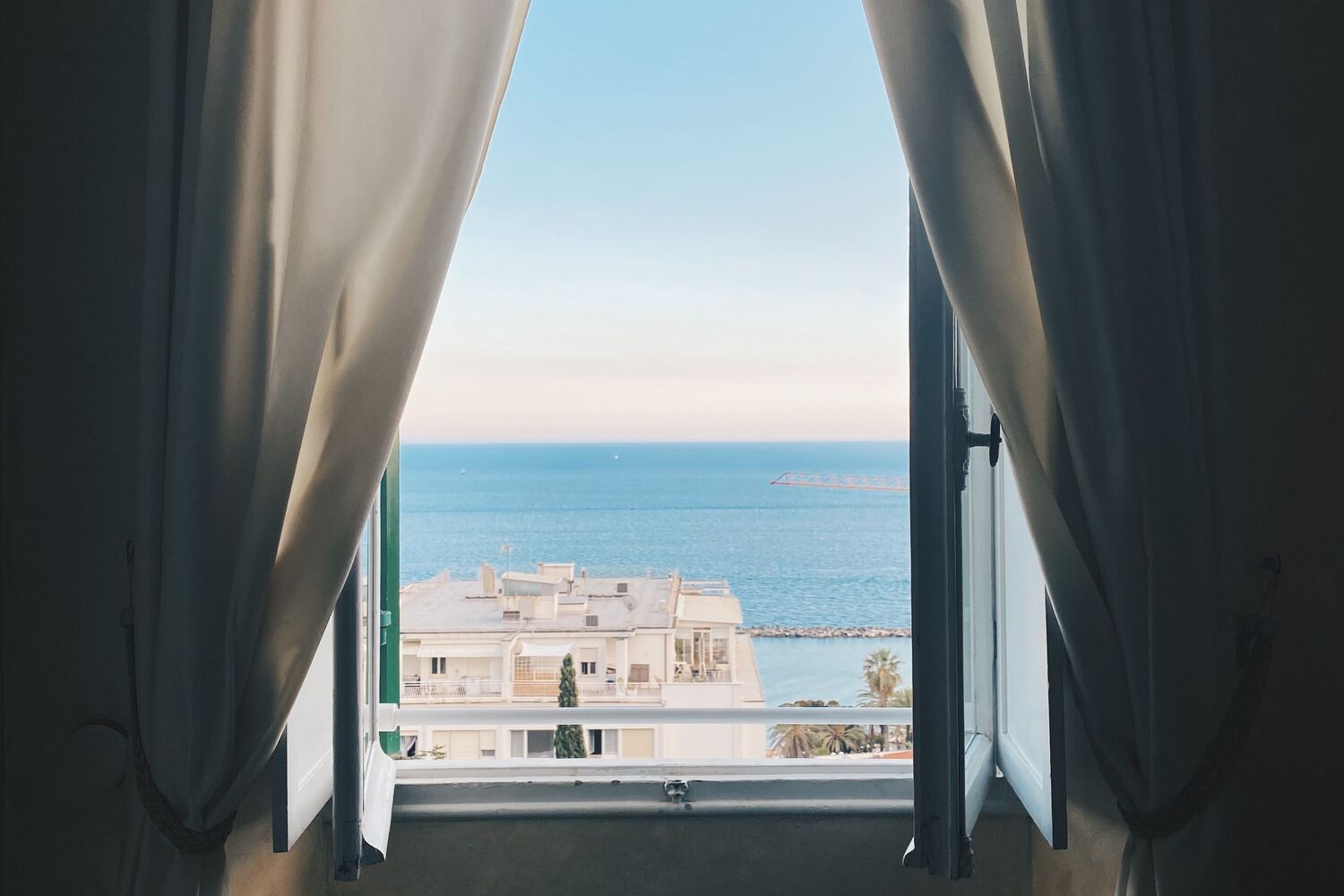 Venezianas abertas e cortinas leves emoldurando a vista do mar sobre prédios brancos baixos