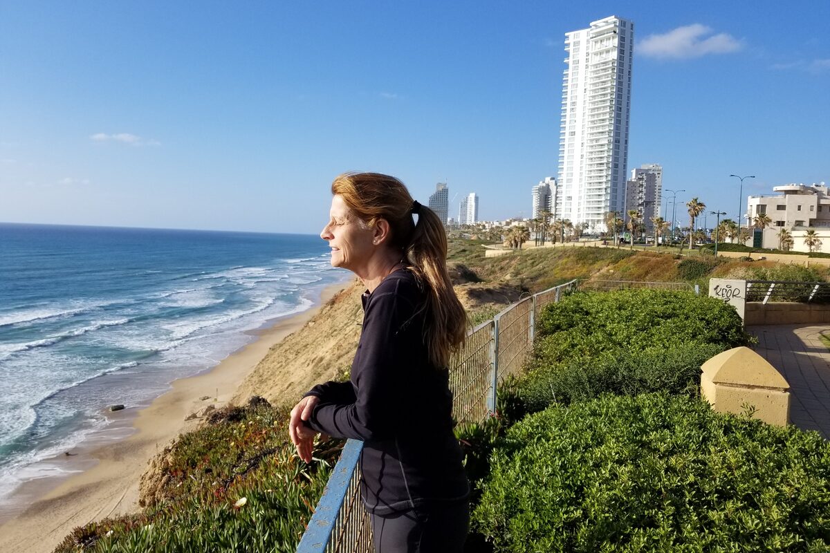 Daniela at a clifftop railing above the sea, the beach below and apartment towers along the shore behind her