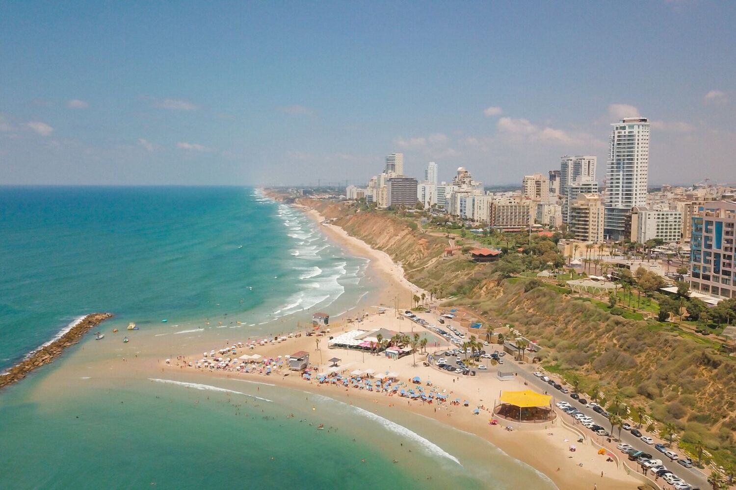 A Mediterranean coastline seen from above: a low cliff, beaches and breakwaters below, and towers along the ridge